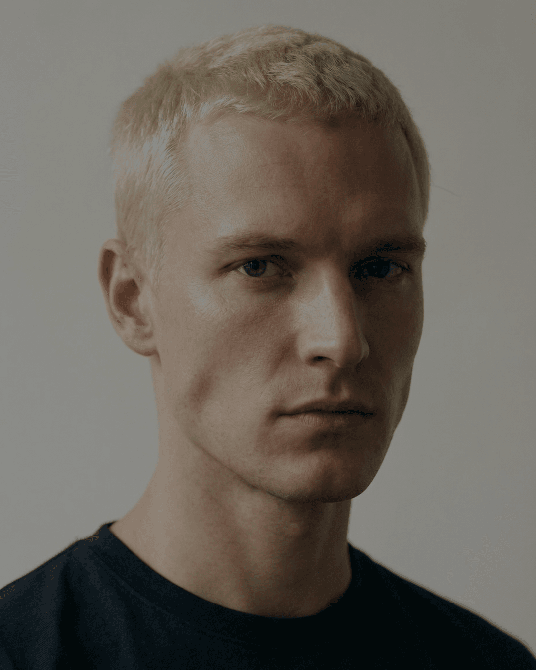 Close-up studio portrait of a model with cropped bleached-blond hair in a black t-shirt, evenly lit against a pale grey backdrop